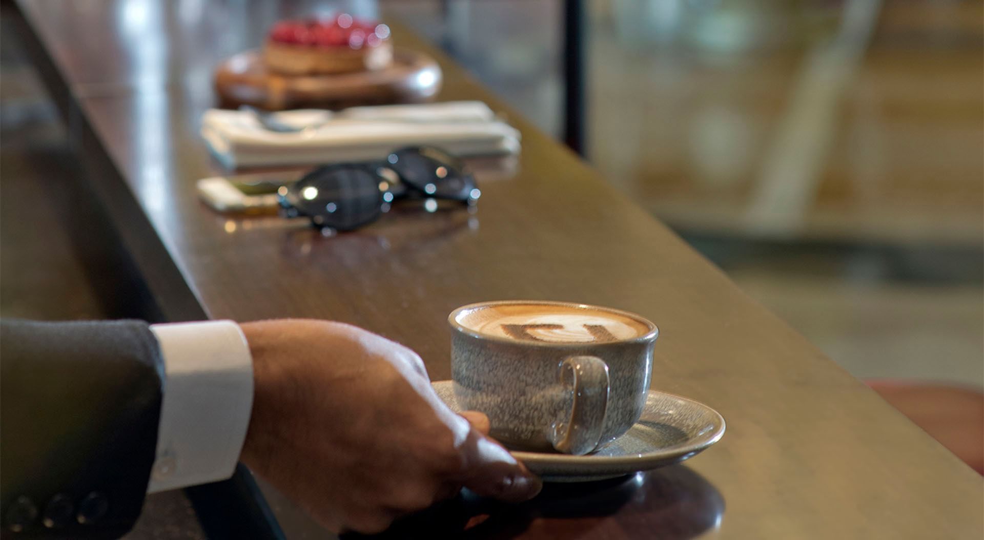 A person's hand in a suit holding a cup of cappuccino on a saucer at a cafe counter at DAMAC Maison Cour Jardin