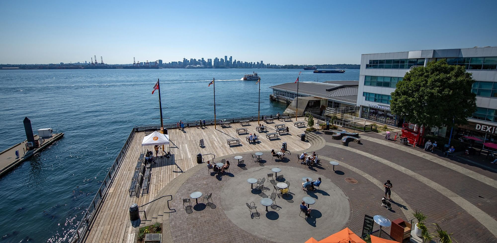 Waterfront patio at Coast Lonsdale Quay Hotel with picnic tables, a large deck, and stunning views of the city skyline