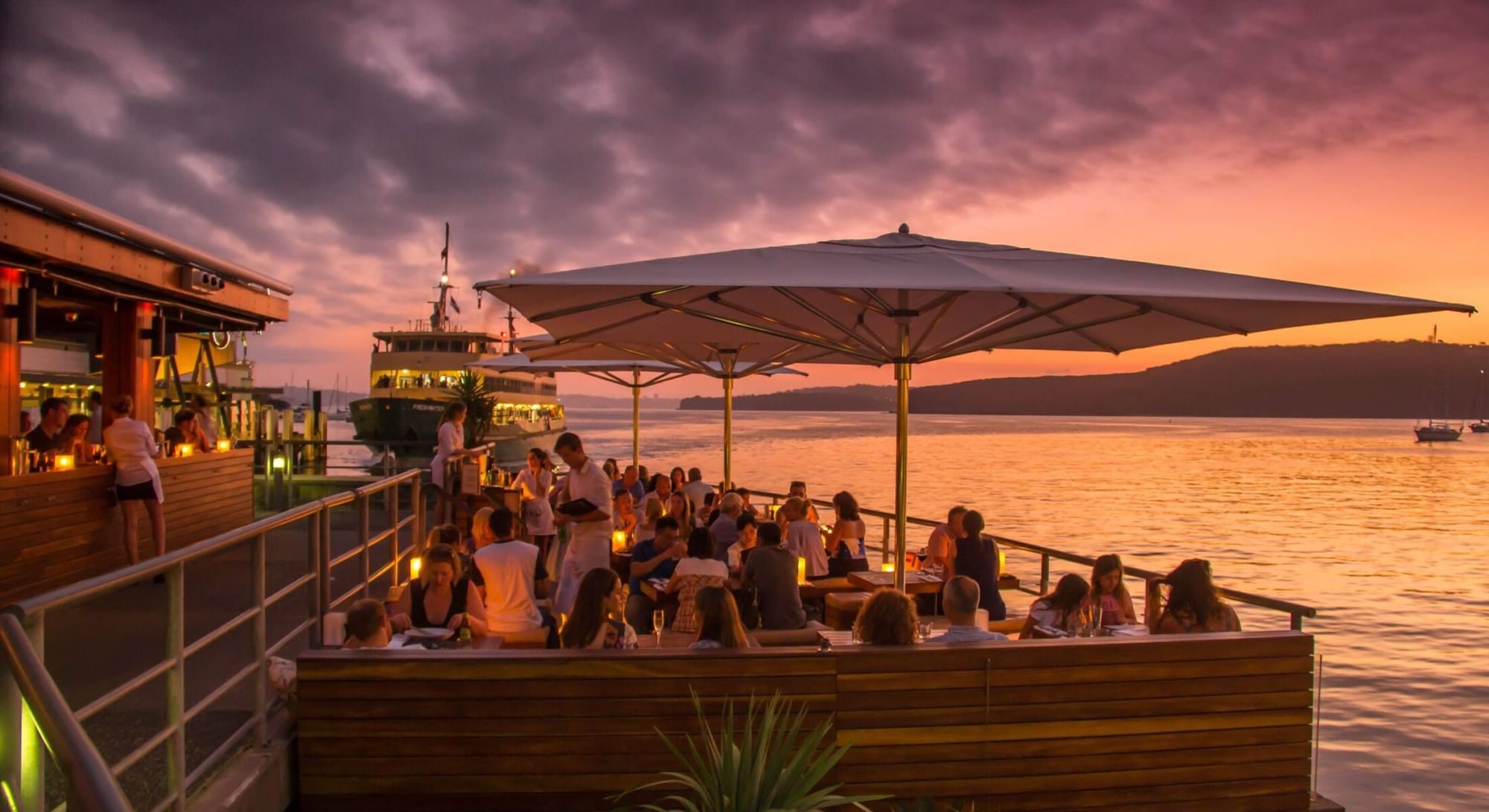 Hugos Manly at Elysium Hotels offers waterfront dining on a wooden deck with a ferry and ocean at sunset