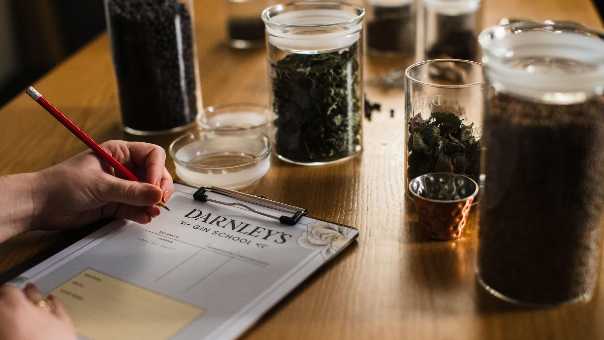 A person writing on Darnley's Gin School clipboard, by the jars of botanicals at Seaton House