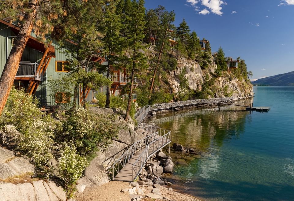 Modern lakeside cabins on cliffside with a metal bridge leading to a dock over clear water.
