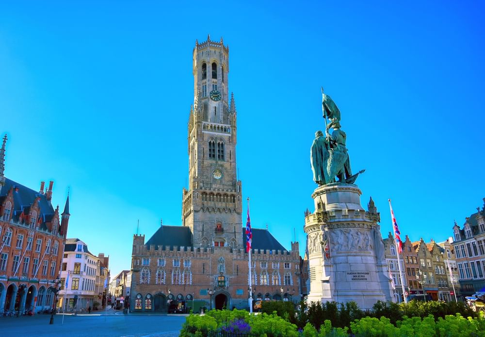 Tall historic clock tower rising behind a stone sculpture, surrounded by square buildings near Warwick Grand Place Brussels
