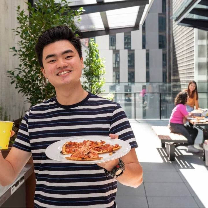 Resident in a striped shirt holds a plate of pizza and a yellow cup outside on terrace.