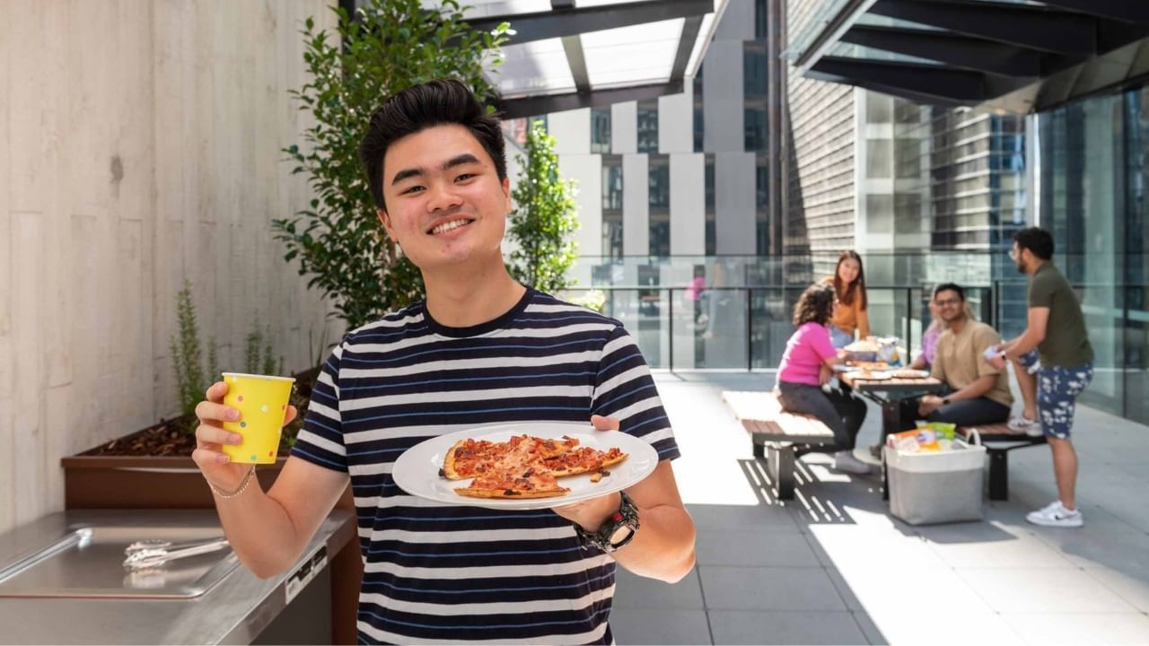 Resident in a striped shirt holds a plate of pizza and a yellow cup outside on terrace.