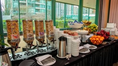 Buffet with cereal dispensers, fresh fruit, pastries, and coffee set beside large windows at Rosedale on Robson Suite Hotel