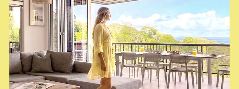 Woman in yellow dress on balcony overlooking ocean with outdoor dining table and cozy living room interior