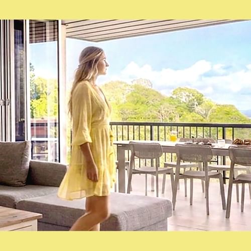 Woman in yellow dress on balcony overlooking ocean with outdoor dining table and cozy living room interior