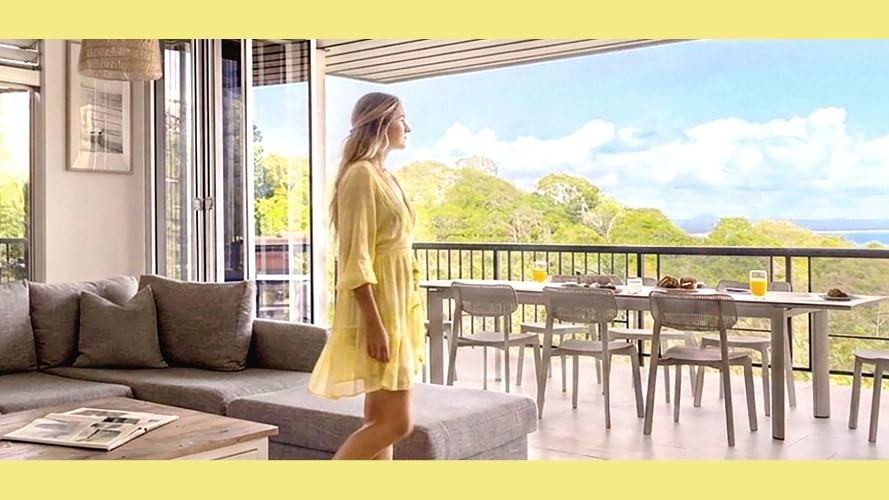Woman in yellow dress on balcony overlooking ocean with outdoor dining table and cozy living room interior