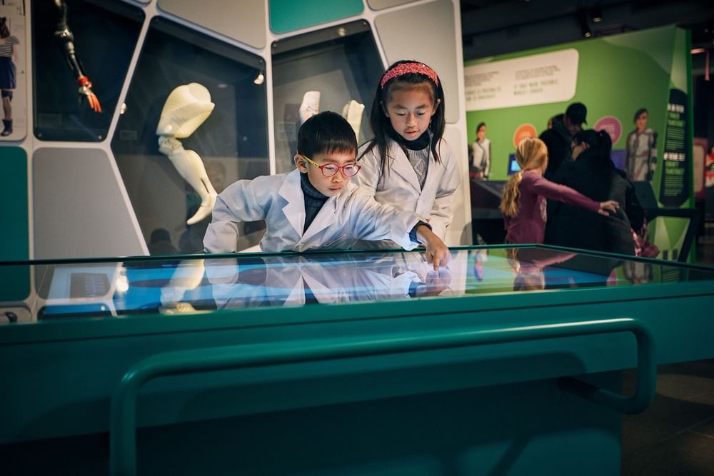 Two children in white lab coats explore an interactive science exhibit at Warwick Le Crystal Montreal