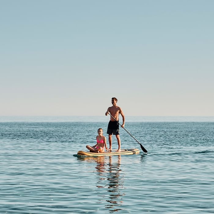 Mann und Frau auf einem Paddelboard im Meer, Mann steht, Frau sitzt, beide halten Paddel.