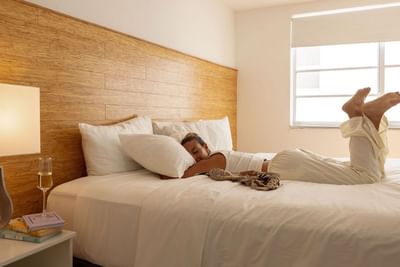 Guest relaxing on a large white bed under a wood headboard by a glass window inside Aqua Hotel and Suites