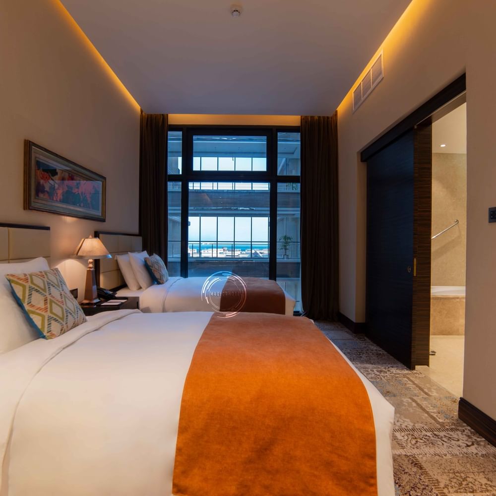 Two twin beds by a lamp under a window overlooking a balcony in Superior Twin Room at Warwick Al Khobar