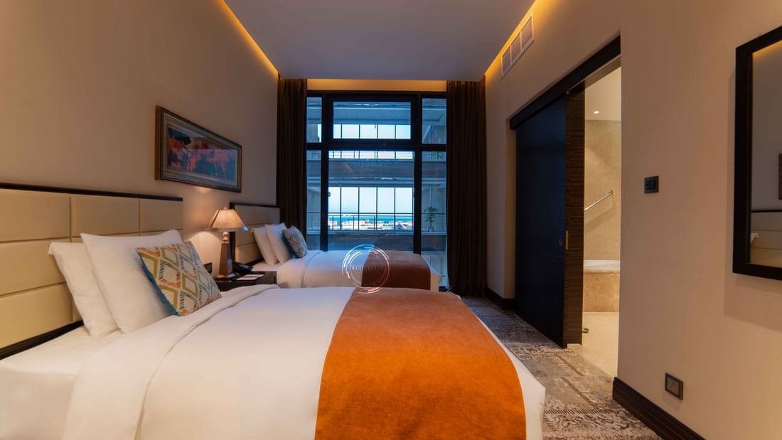 Two twin beds by a lamp under a window overlooking a balcony in Superior Twin Room at Warwick Al Khobar