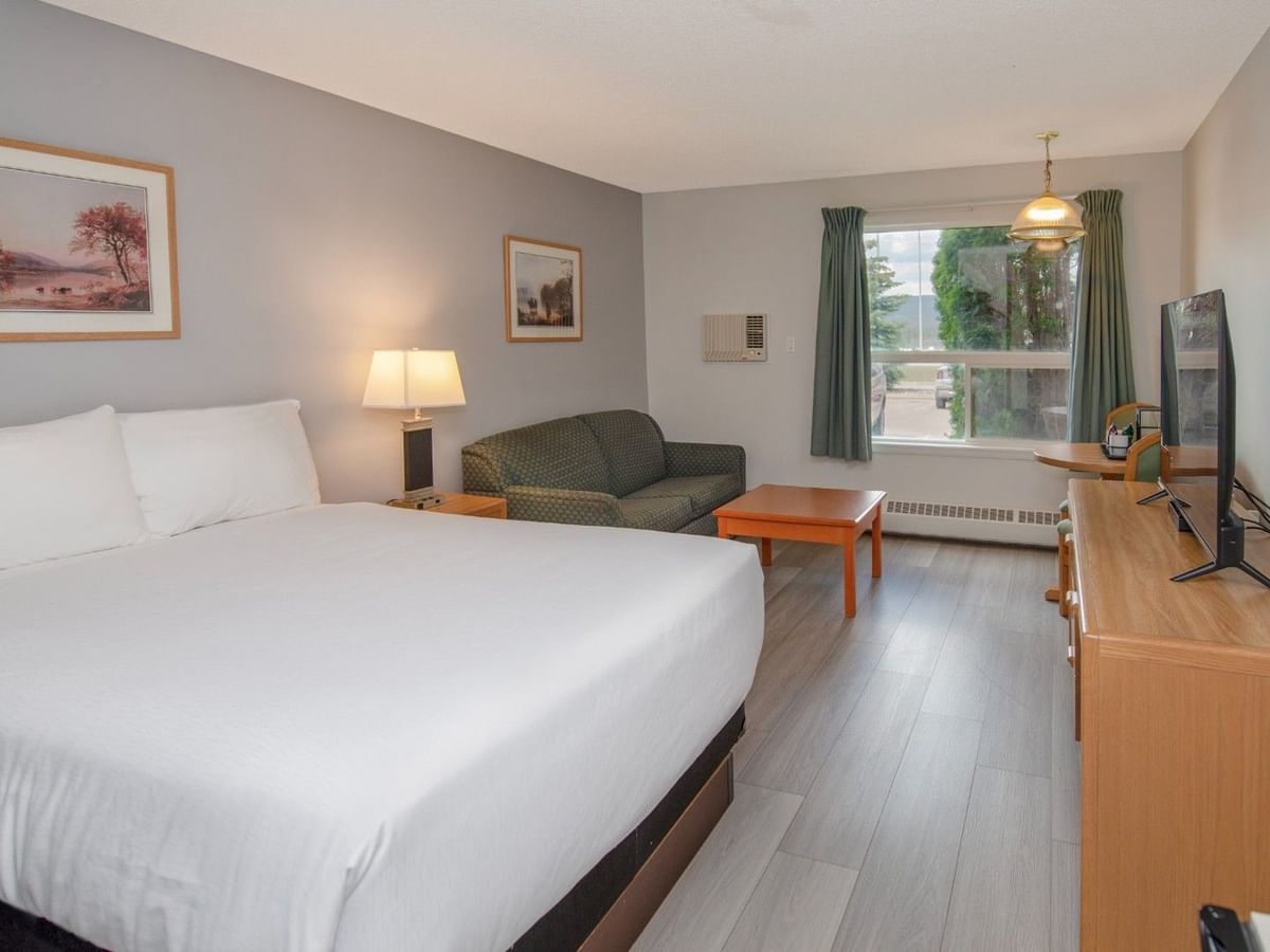 A hotel room with a white bed, green couch, wooden furniture, TV, and artwork.