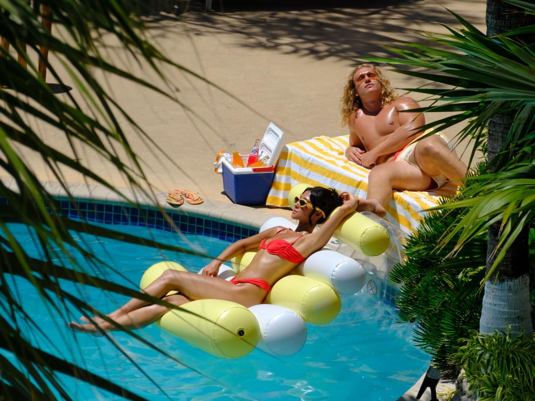 Couple relaxing by the pool at Tradewinds Apartment Hotel, with one guest floating in the water and the other sunbathing