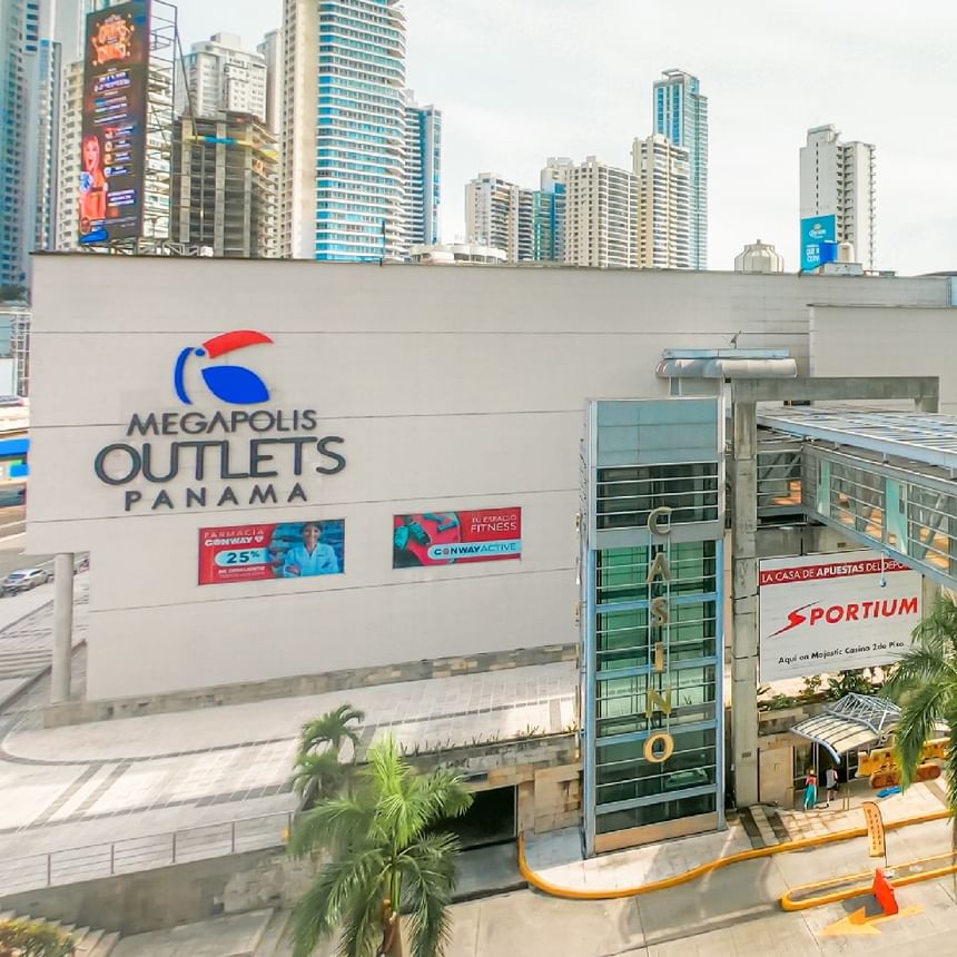 Exterior de Megapolis Outlets Panama con entrada de vidrio, palmeras y rascacielos cerca de Megapolis Hotel Panama