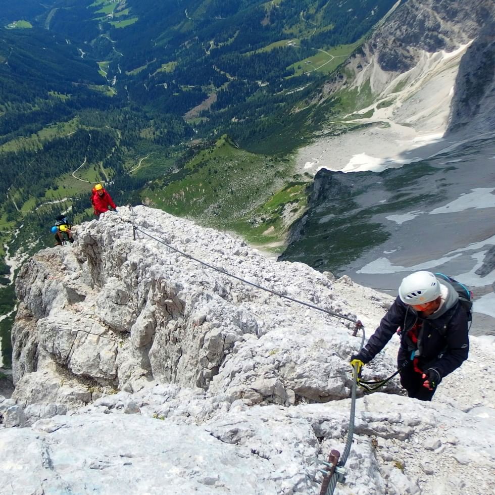 Kletterer mit Helm und Sicherheitsausrüstung auf einem alpinen Klettersteig mit Stahlseil und Bergpanorama im Hintergrund