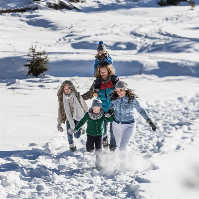 Family of five in winter clothes walking in snow, two kids on parents' shoulders.