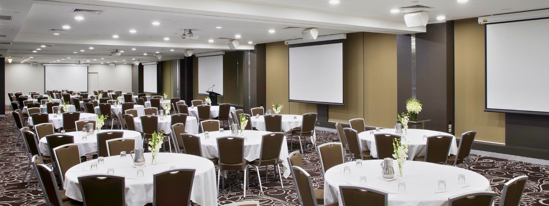 Large conference room with round tables, white tablecloths, brown chairs, and three large white projection screens on walls.
