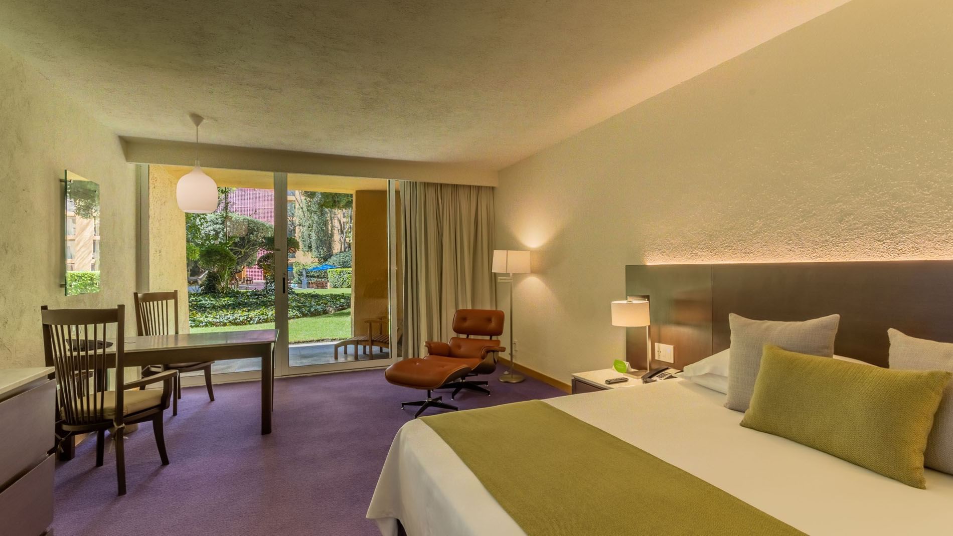 Habitación de hotel con cama grande, sillas, mesa, lámparas y balcón con vista al jardín.