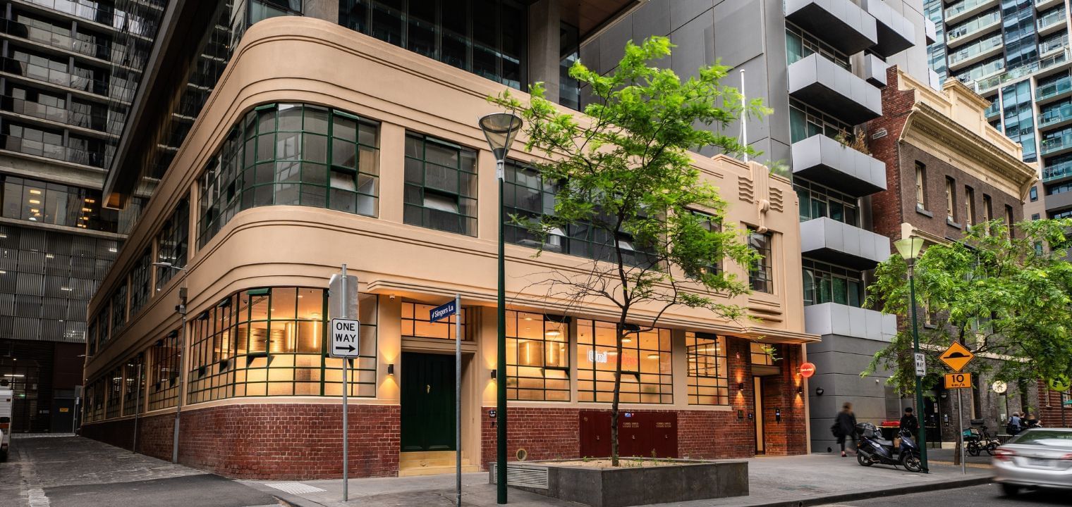A beige UniLodge building with green windows and a tree in front, next to a road with cars and scooters.