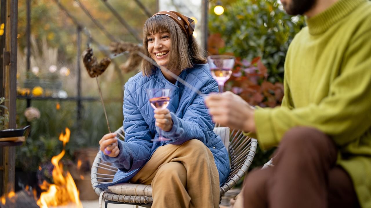 Woman in blue jacket roasting marshmallow over fire pit while holding wine glass.