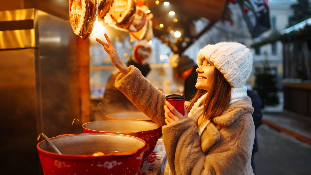 A smiling woman in winter attire points upwards while holding a red cup at a holiday market.