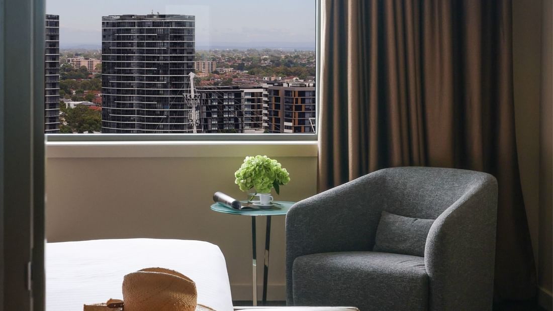 Modern hotel room with gray armchair, side table with flowers, and panoramic city view through large window.