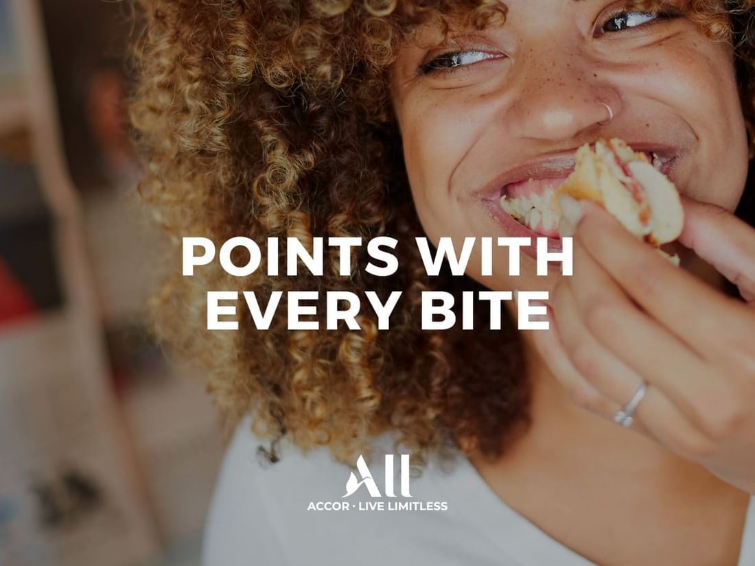 Joyful woman with curly hair takes a bite, highlighting earning Points With Every Bite at The Sebel Mandurah