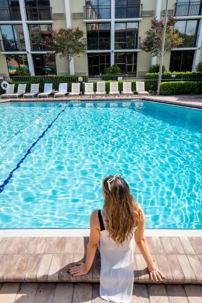 A woman in a white bathing suit cover sits on the edge of a sparkling blue pool with lounge chairs and a hotel building in the background.