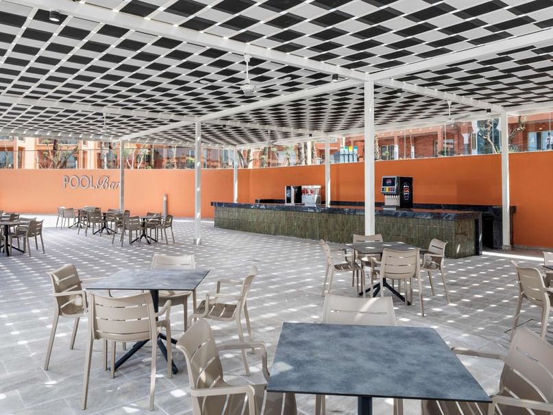 Outdoor seating area with tables and chairs under a patterned roof, and a bar counter in the background.