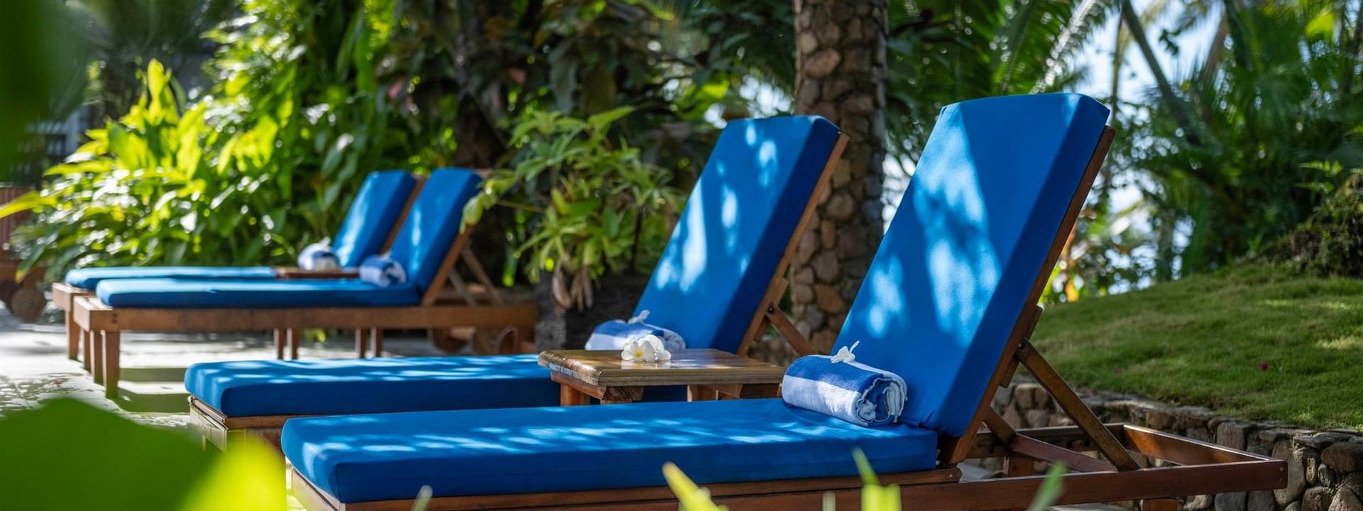 Blue sun loungers by wooden tables under leafy palm trees surrounding a garden at Warwick Fiji Resort and Spa Korolevu