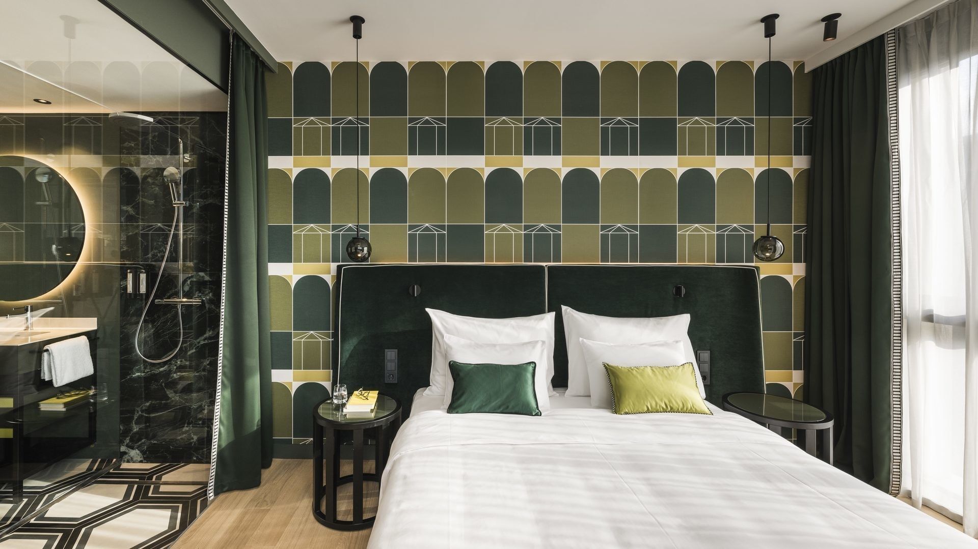 Modern bedroom with patterned wallpaper, green velvet headboard, white bed, and adjacent bathroom.
