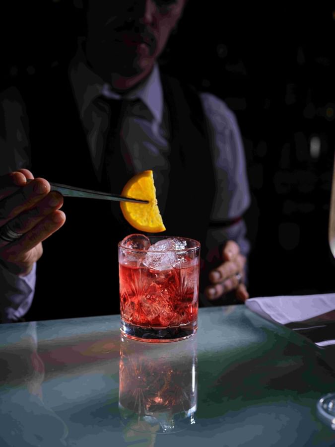 Bartender using tweezers by a bar to place an orange slice in a red cocktail at The Capital Hotel, Apartments and Townhouse
