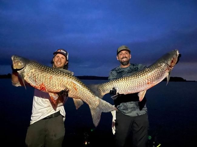 Two smiling men hold up two very large, heavy fish they caught at night near a lake at Shangri-La Resort and Golf Club
