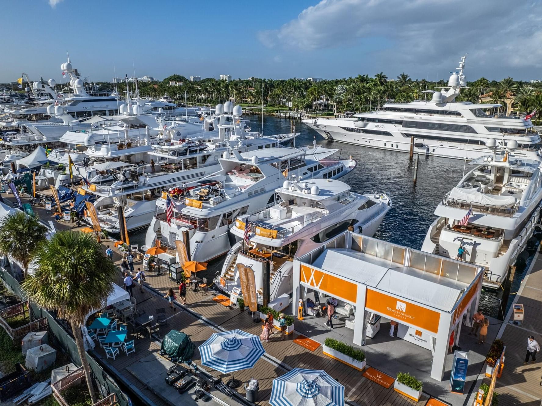 Aerial view of luxury yachts and superyachts docked at a waterfront marina with palm trees and visitors exploring the displays