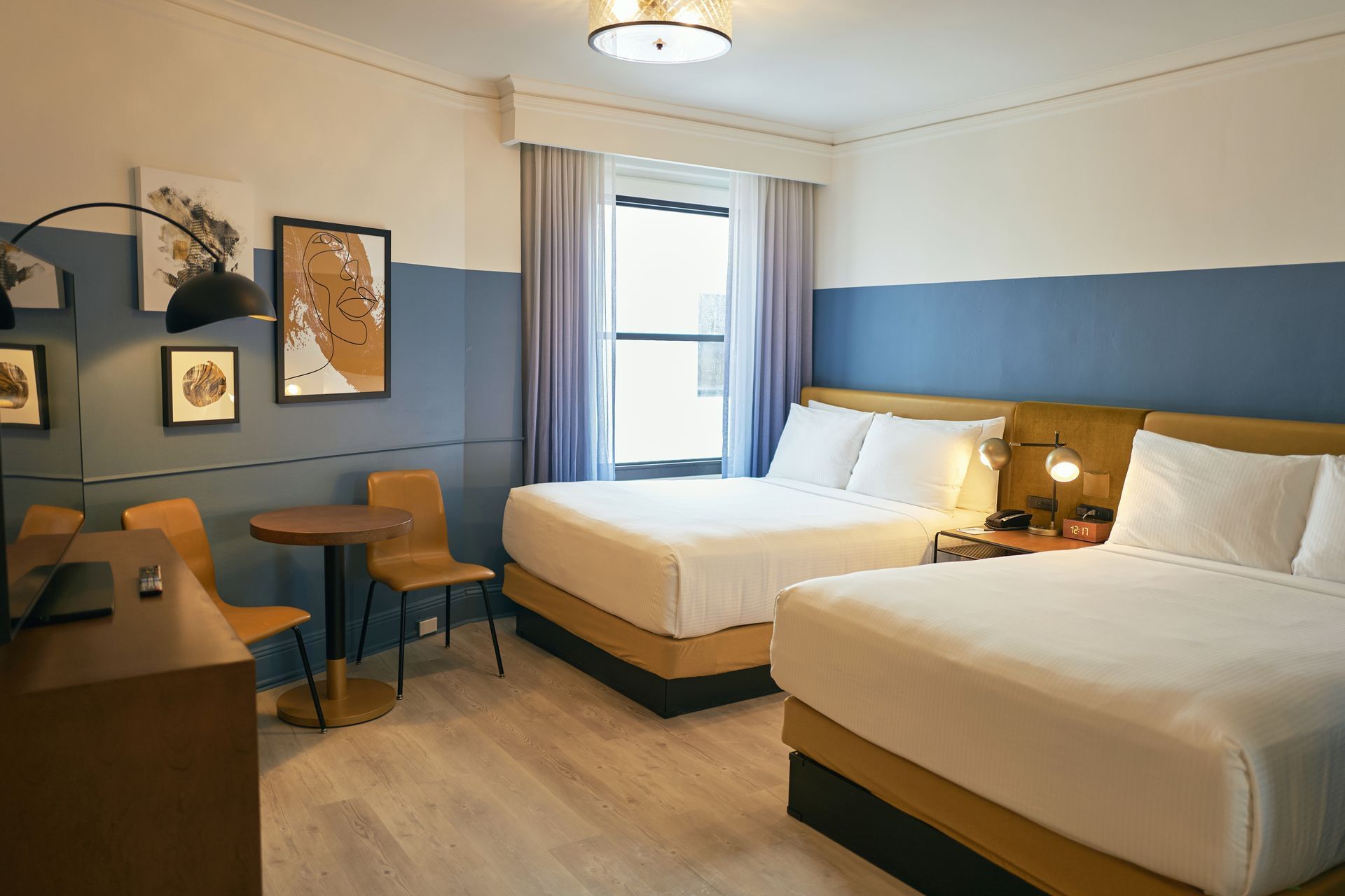 A hotel room with two queen beds, a desk, table and chairs, and artwork on the walls.