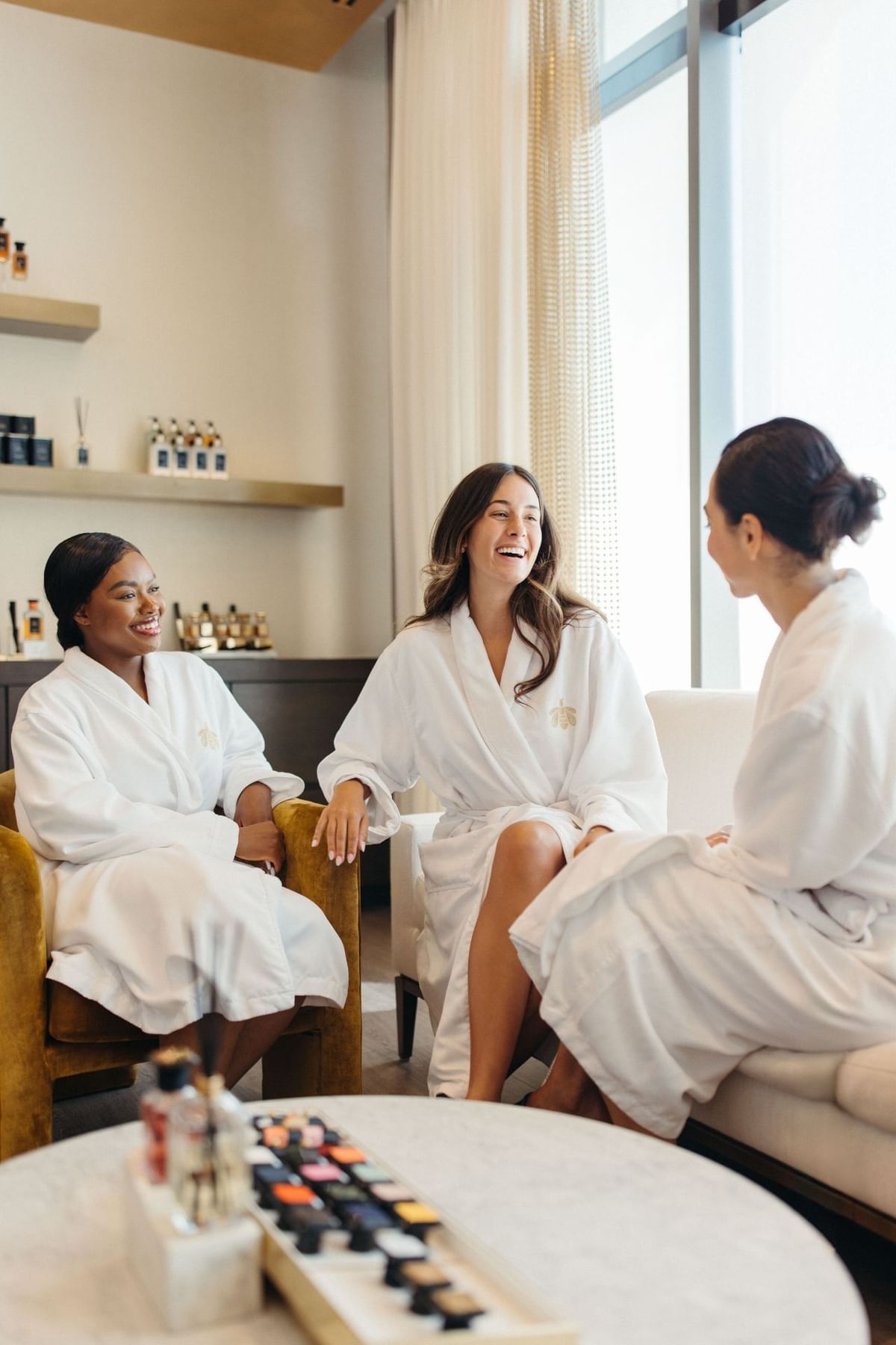 Three smiling women in white bathrobes chatting on armchairs inside a luxury spa lounge area at Hotel X Toronto