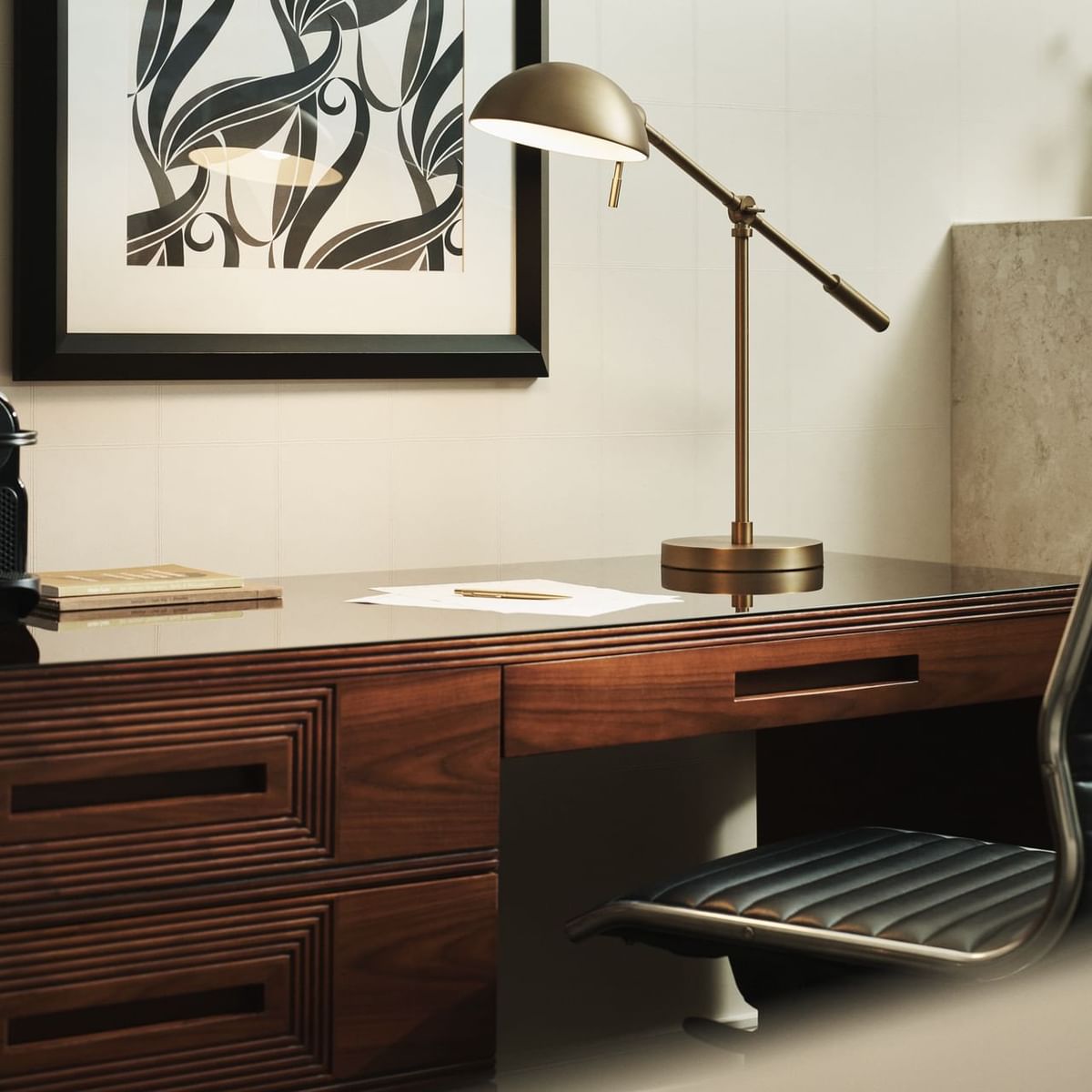 A desk with a lamp, chair, coffee maker, book, cup, and a framed print on the wall.
