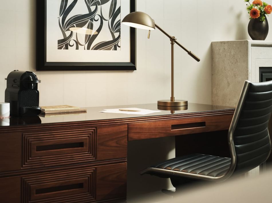 A desk with a lamp, chair, coffee maker, book, cup, and a framed print on the wall.
