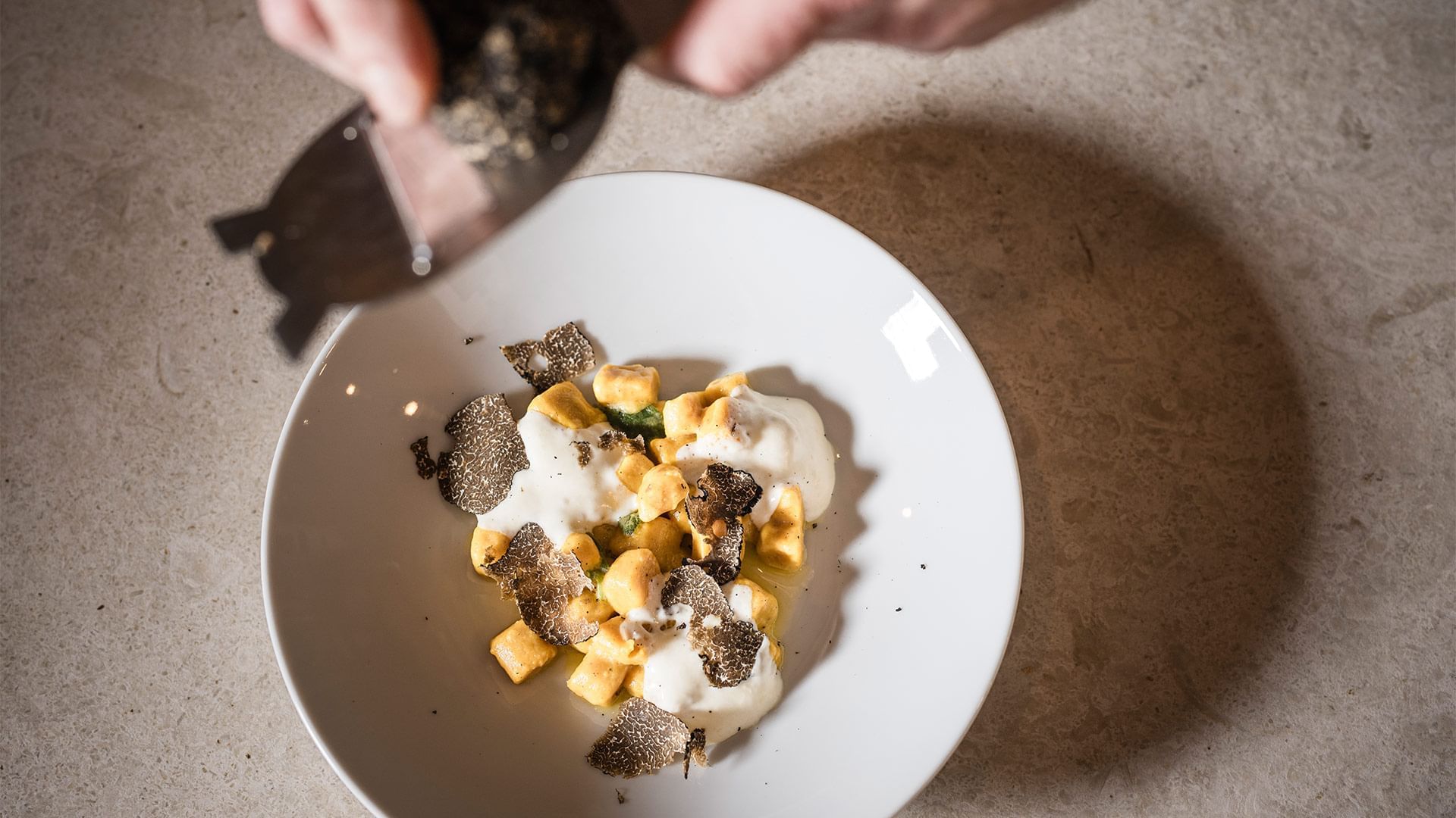Mano aggiungendo tartufo nero su pasta gialla con crema bianca in un piatto bianco su superficie chiara.