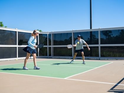 Two people playing pickleball on an outdoor court with a mesh fence and palm trees in the background.