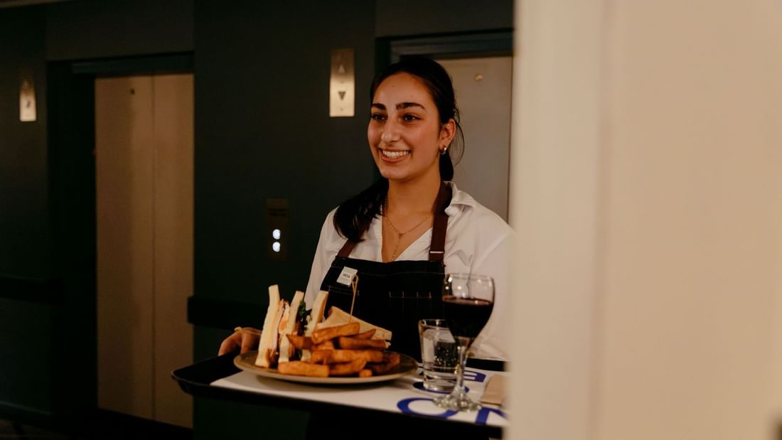 A smiling waitress holds a tray with a sandwich, fries, and a glass of wine.
