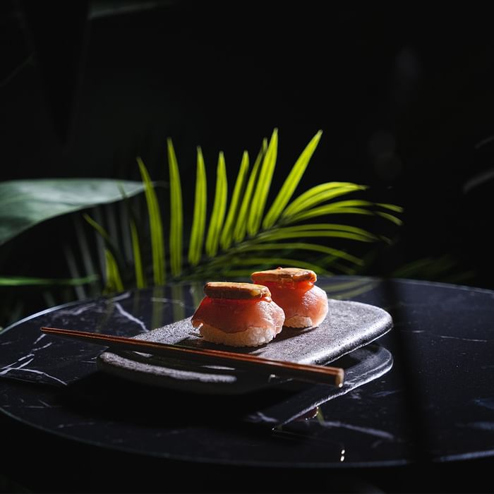 Two sushi rolls on a dark plate with chopsticks, set against a leafy background.