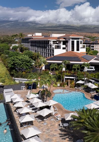 Exterior of Maui Coast Hotel, highlighting the lounge chairs with umbrellas by the pool and mountains in the background