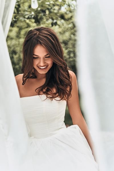 Smiling bride with long hair wearing a strapless gown surrounded by white curtains at Spencer Hotel & Conference Centre