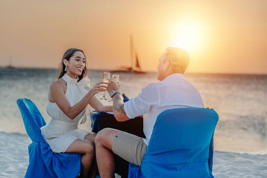 A couple at Passions on the Beach at Eagle Beach Aruba toasting with wine glasses at a beach sunset, seated on blue chairs.