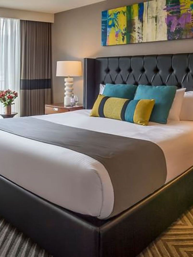 King bed with a black headboard by a colorful painting and city view windows in Emerald Suite at Warwick Seattle