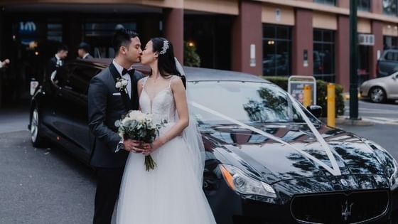 Stunning wedding couple kisses beside a black luxury car with white ribbons near Amora Hotel Riverwalk Melbourne