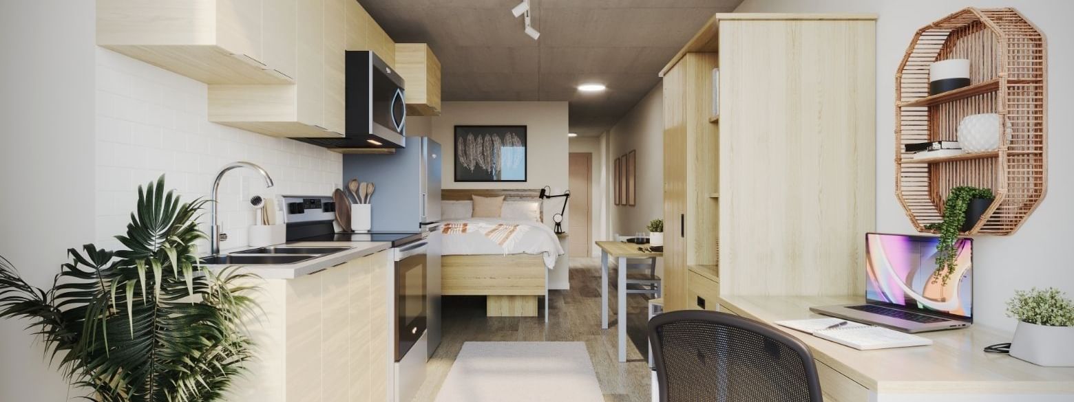 A large and bright studio apartment containing a bed, a study area, and a full sized kitchen.